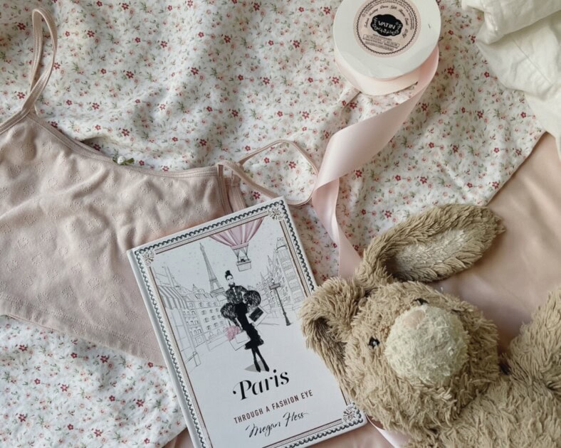 Flatlay photograph with a stuffed bunny, pink crop top, pink satin ribbon, and fashion book about Paris on a floral pink background, a perfect coquette look inspired by Redfin's ultimate coquette room decor guide